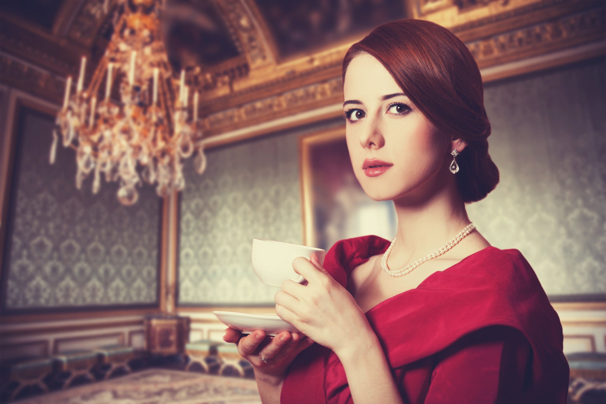 A woman in a red dress, adorned with delicate jewellery, holds a teacup and saucer in an ornate, chandelier-lit room with decorative Edwardian walls.