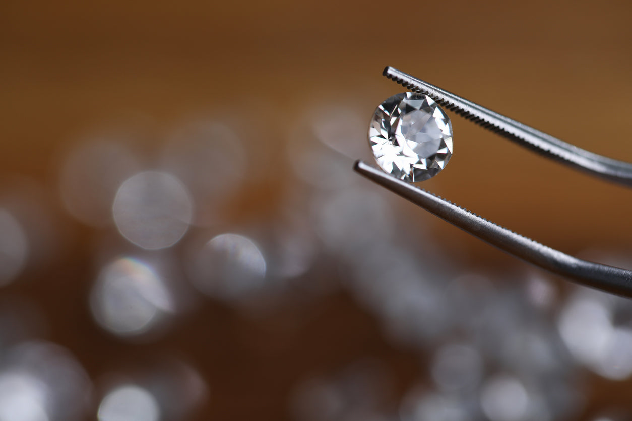 Painstaking jewelry work, looking at diamond. Master holds small gem with tweezers. In foreground, this object with many faces shimmers with natural light, and other pebbles laid out on table.