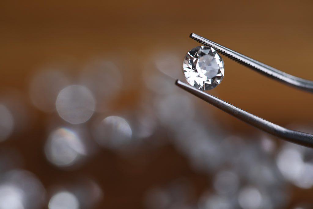 Painstaking jewelry work, looking at diamond. Master holds small gem with tweezers. In foreground, this object with many faces shimmers with natural light, and other pebbles laid out on table.