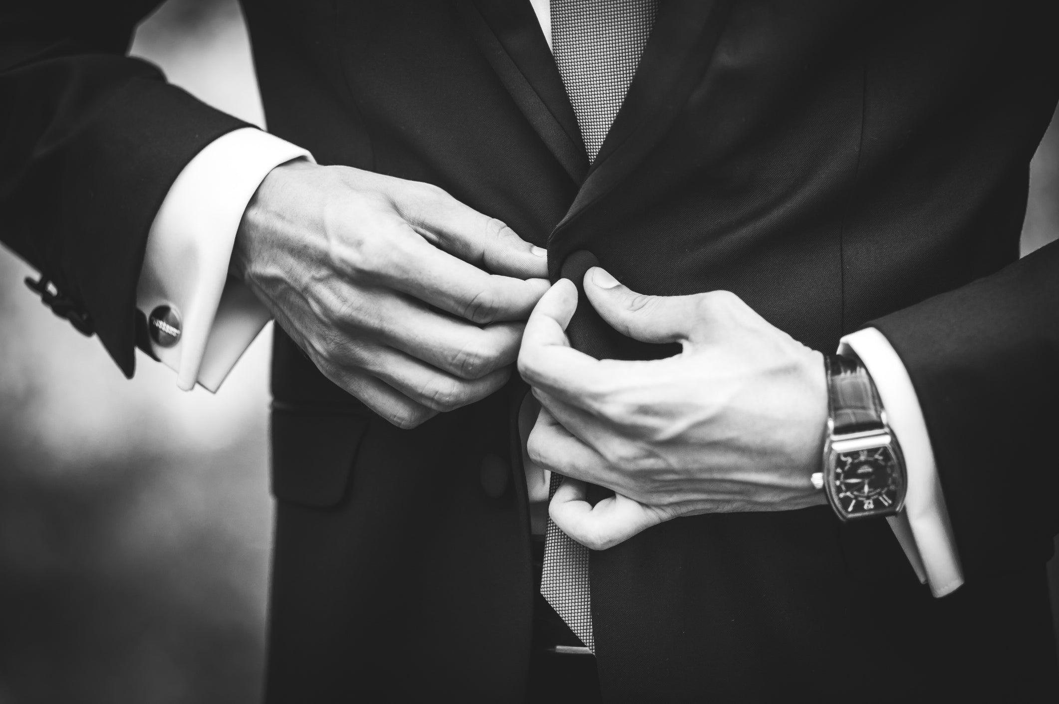 A black and white photo of a man buttoning his suit jacket, his cufflinks visible